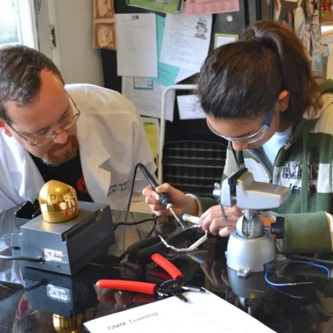 At site C, a girl follows the adult educator's instructions to solder two wires together to make a bracelet. In this participation structure, known as scripted build, the educators give young people instructions and monitor their progress. This structure was introduced by the educators and was not part of the 4-H curriculum.