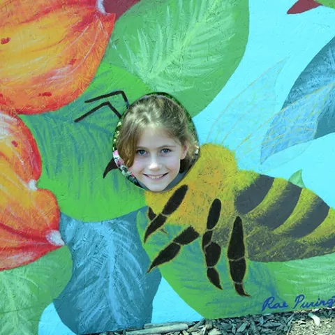 Beekeeper Adelaide Grandia smiles through a pollinator cut-out board. Her grandfather is teaching her beekeeping. (Photo by Kathy Keatley Garvey)