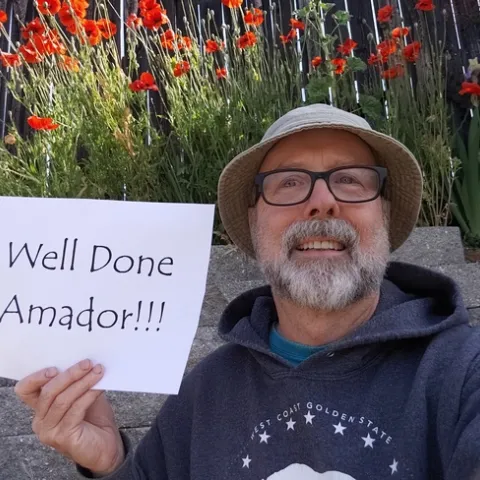 Photo of UC Master Gardener volunteer Ed Bass wearing a gardening hat and glasses, holding up a sign in his gardener reading "Well Done Amador"