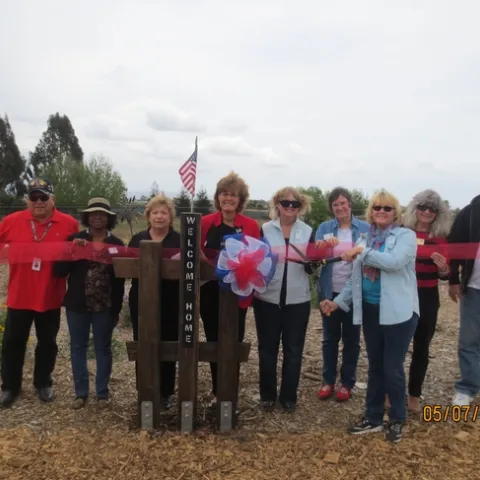 Nine people stand on a wood chip covered garden path, behind a post, engraved vertically with the words welcome home.” A small American flag is mounted atop the post. One person holds garden shears, poised to cut through a translucent red ribbon being held by eight other gardeners.