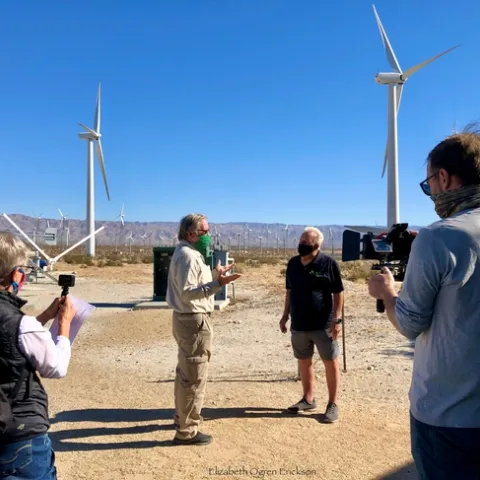 With everyone wearing face masks, the four people stand on dusty soil among dozens of three-bladed windmills, under a cloudless blue sky.