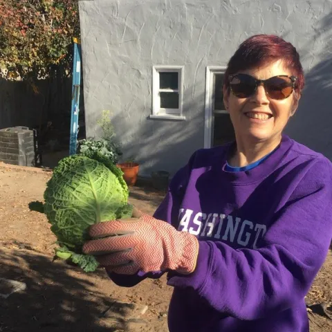 Elaine Silver, wearing sunglasses and a purple sweatshirt with Washington printed in white, holds up a green head of cabbage.