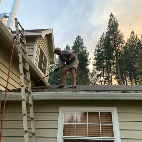 Ryan Tompkins stands on his roof raking moss, vegetative debris, and leaf litter from the gutters.