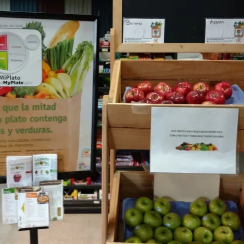 Baskets of apples in a small market are displayed along with Spanish and English language signs encouraging customers to eat fruits and vegetables