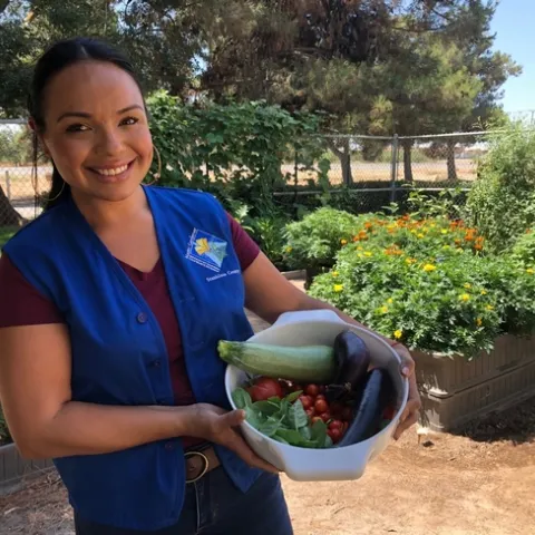 Master Gardener smiling with basket of garden harvest of eggplant, squash, basil, and tomatoes.