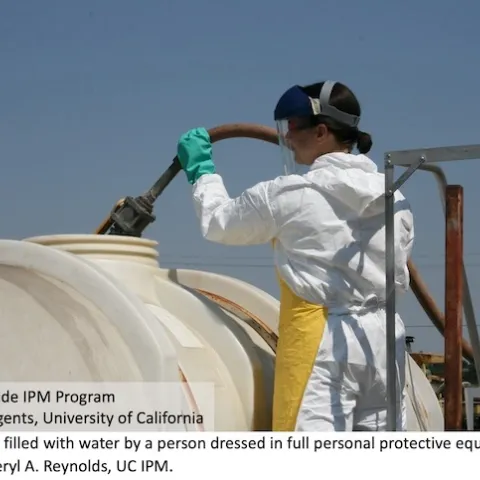 A person filling a tank with water while wearing chemical-resistant gloves, coveralls, chemical-resistant apron, and a face shield over safety glasses.