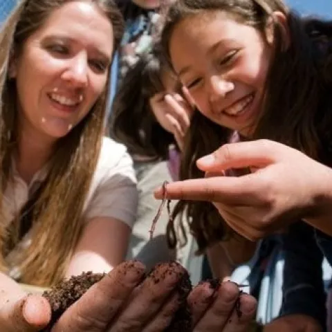Woman holding handful of worms and compost while teaching a smiling young girl.