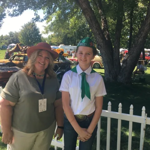 June, wearing a hat, stands under a leafy tree beside Lily, who is wearing a 4-H uniform of green and white. Classic cars are in the background.
