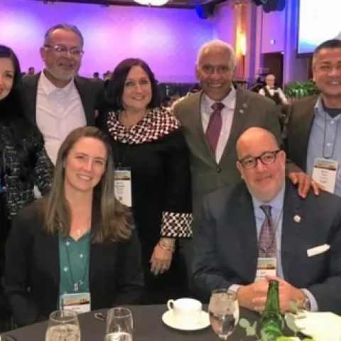 Nine people - 2 sitting and 7 standing - around a table in a conference hall.