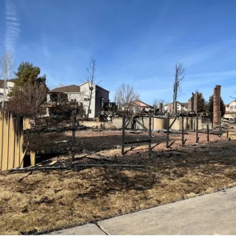 Fire scorched grass black in the foreground. A few yellow boards of the fence remain standing beside the burned posts.