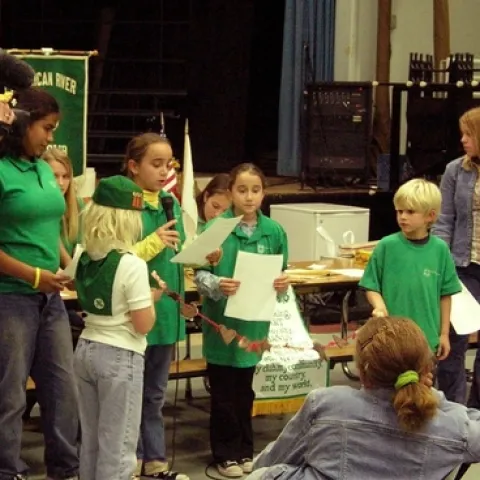 Kids in green shirts read from pieces of paper as someone shoots video.