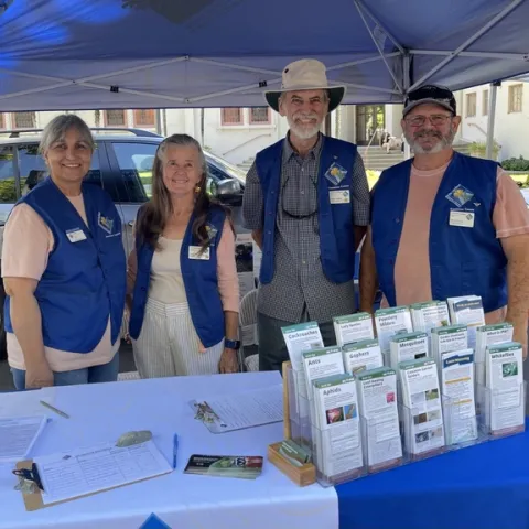 Four smiling people wearing blue vests behind a table with brochures.