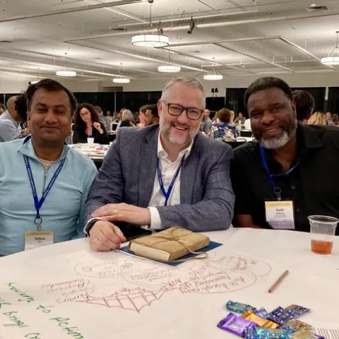 Three smiling men sit at a round table in a conference hall.