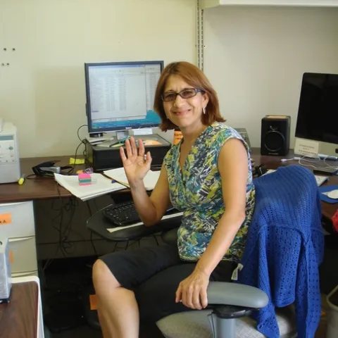 A woman sits at a computer, in a chair, waving at the camera.