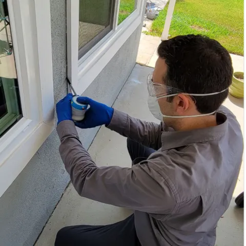 A man wearing a mask, gloves, long sleeve shirt, and pants applying a pesticide dust into a crack on an exterior wall.