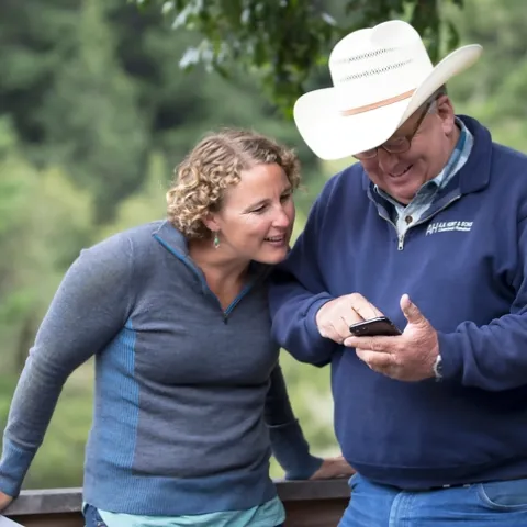 Lenya smiles and looks at whatever a man in a cowboy hat is pointing at on his phone.