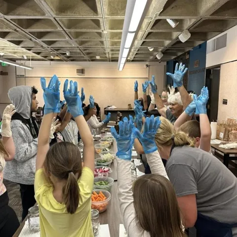 A table set-up with food and youth with gloves on and hands in the air. Adults providing instructions
