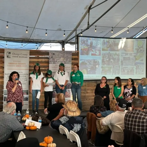 A woman speaking into a mic introduces the 4-H members, who are wearing green and white uniforms standing on a stage. Audience members sit in the foreground.