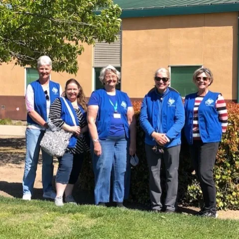 Five smiling women in front of a garden wearing blue vests.
