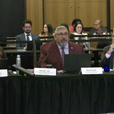 Three people sit at a long table facing the camera. A gallery of spectators sit behind them.