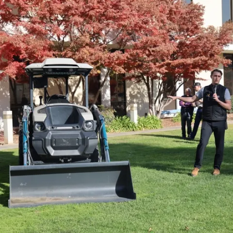 Standing on a lawn, a man gestures toward a piece of machinery.
