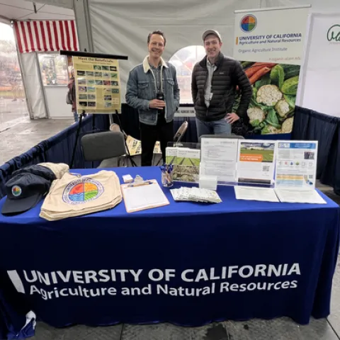 The two men are standing behind a table of OAI logo hats and bags and brochures.