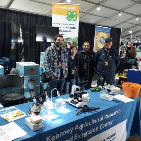 Four people stand behind a table covered with a blue tablecloth labeled Kearney Agricultural Research Extension Center and next to boxes labeled RECs