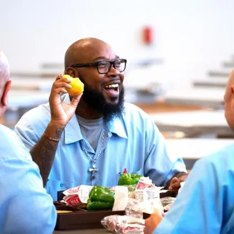 Three bald men wearing blue prison shirts sit at a table eating lunch.