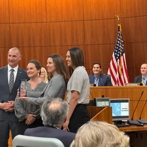 Four people pose for a photo with Shannon holding up the lucite trophy as the board of supervisors look on in the background.