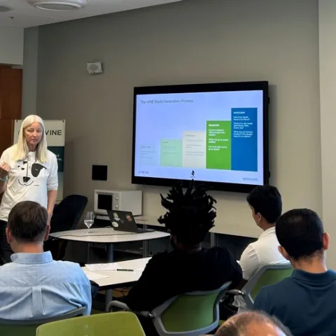 A woman speaks to a group of people sitting at desks. The slide on the screen reads: The VINE Studio Innovation Process.