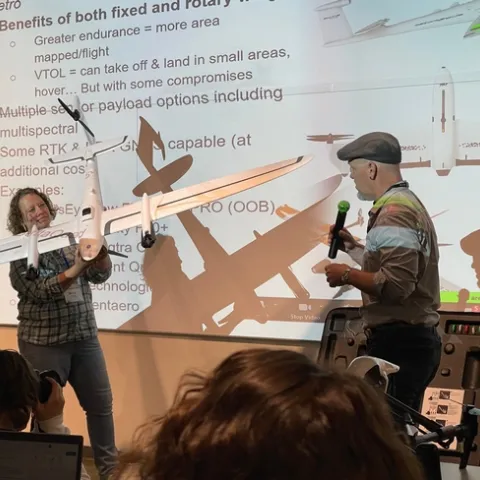 Becca holds a large model airplane as a man wearing a hat speaks into a handheld microphone.
