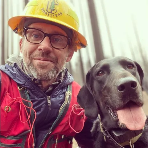 Ryan, wearing a red jacket and yellow hard hat that reads, "Cal Foresters" around a C with an evergreen tree in the middle, poses beside his black labrador, whose pink tongue is hanging out.