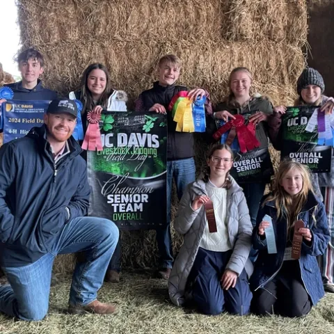 Against a backdrop of hay bales, 5 youth hold ribbons and posters of their contest placing. Kneeling in front of them is an adult coach and 2 youth holding ribbons.