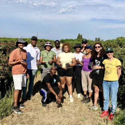 Eleven young people holding clear plastic cups of blackberries pose between rows of blackberry bushes.