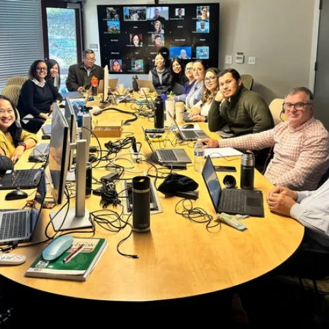 In a conference room, 14 people sit around a conference table littered with laptops.