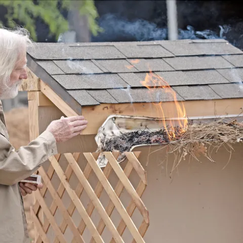 Steve Quarles gestures toward flames rising from dry pine needles and leafy debris in a melting plastic rain gutter affixed to a structure with a shingled roof.