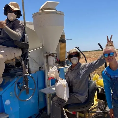 A person sits in the driver's seat of a combine, another person sits beside the combine holding a bag in his right hand while making bunny ears with his left hand behind Maya's head as she stands beside him.