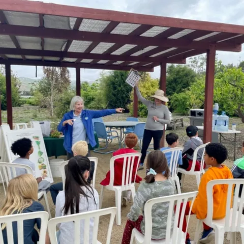 Two middle aged women stand in front of a crowd of seated children, teaching them about food.