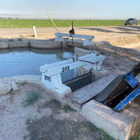 Gates are open, releasing water into an irrigation ditch running between a road and a crop field.