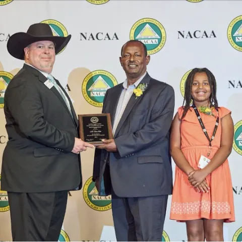 A man in a cowboy hat hands Oli a plaque. Marti stands beside Oli.