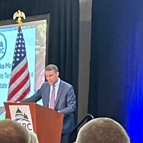 McGuire stands at a lecturn beside an American flag, speaking into a mic.