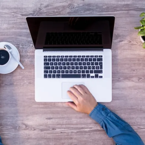 An overhead shot of a person with one hand holding a coffee mug and the other hand on the mousepad of a laptop.