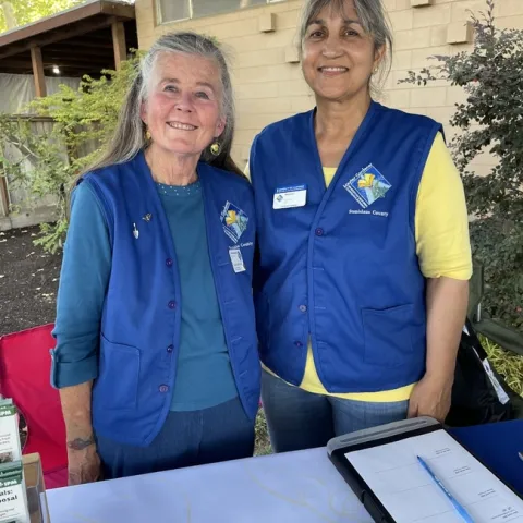 Two women wearing blue vests and smiling while staffing a booth.