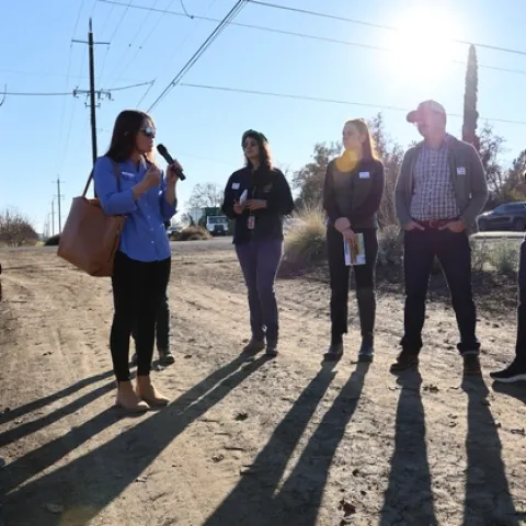 Hope, surrounded by 6 people standing on a dirt road, speaks into a mic.