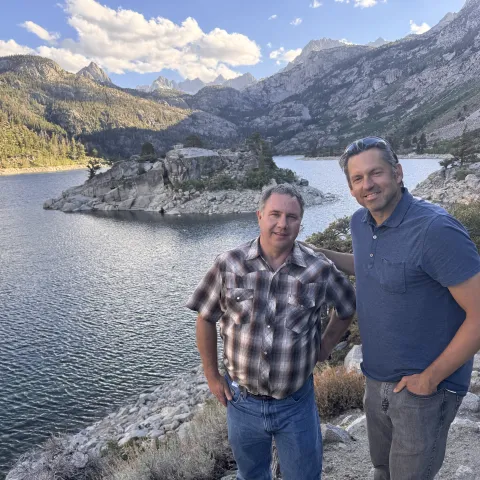 Two men stand at overlook above Sierra lake with peaks in the distance