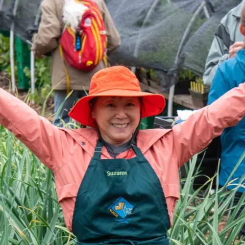 A smiling UC Master Gardener volunteer in an orange hat and green apron stands with arms raised in a garden setting, surrounded by people and plants.