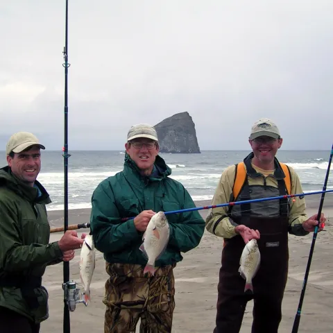 Three men at the beach, each holding fishing poles and a fish