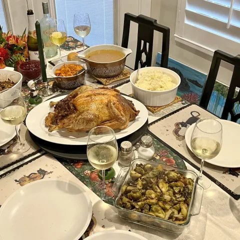 A dining table with stuffing, sweet potatoes, gravy, a roast turkey, mashed potatoes, Brussels sprouts, green bean casserole, and five glasses of wine.