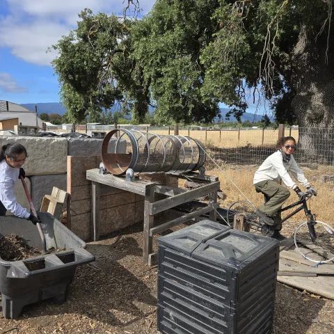 Two people sifting raw compost using a bike propelled trommel screen, one loading with a shovel, the other peddling the bike