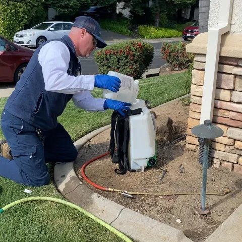 A man kneeling on a lawn next to a house pouring a liquid pesticide into a plastic backpack applicator.